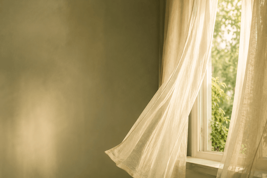 A single softly lit window with sheer curtains gently moving, suggesting a breath of air moving through a calm room